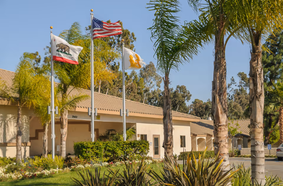 Exterior view of a single-story building with a tiled roof, surrounded by palm trees and other greenery. Three flagpoles display the California state flag, the American flag, and another flag with a yellow emblem. The sky is clear and blue.