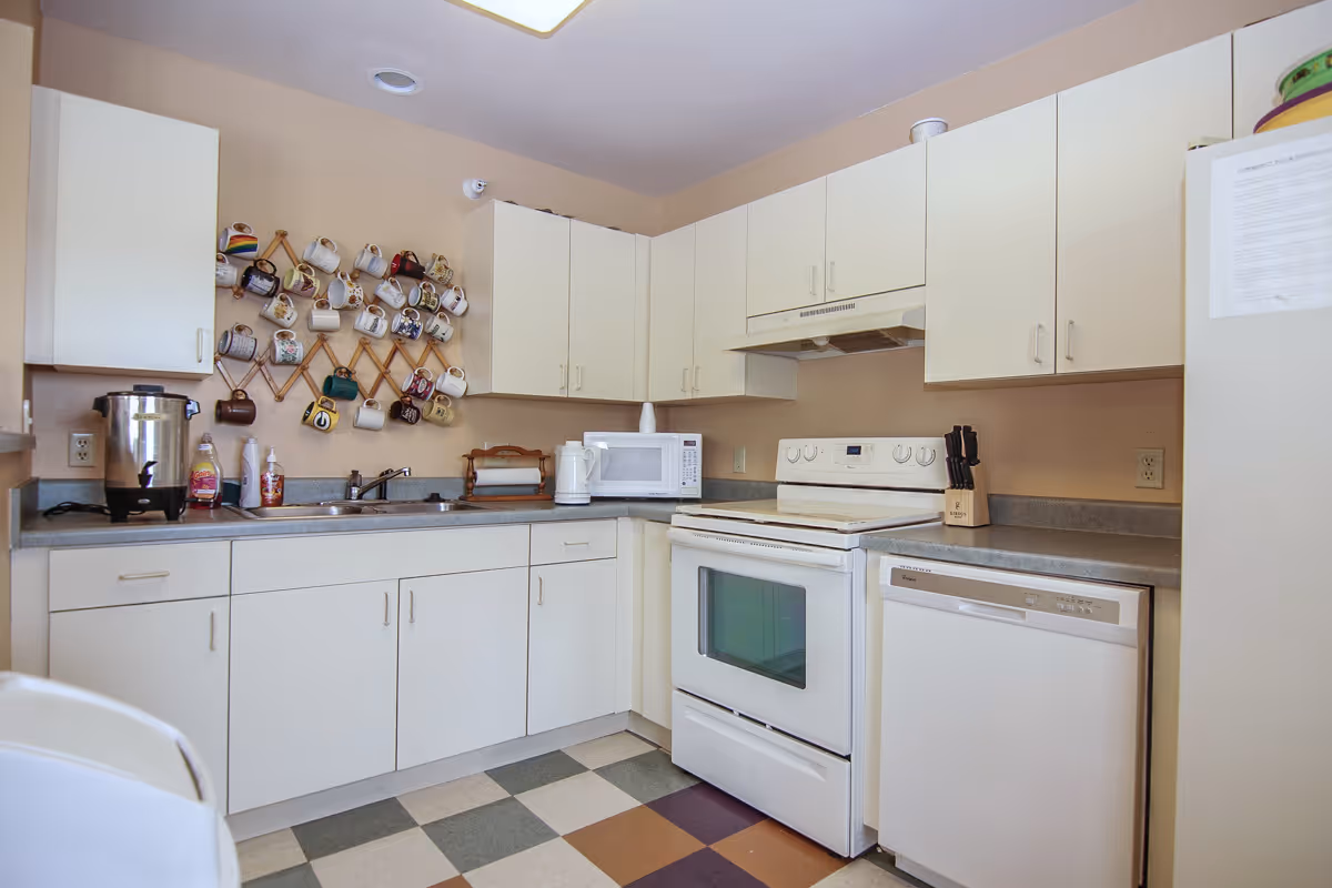 A kitchen with white cabinets and appliances including a stove, microwave, dishwasher, and refrigerator. There is a countertop with a sink, a coffee urn, soap dispenser, and a rack on the wall holding various coffee mugs. The floor has a checkered pattern with different colored tiles.