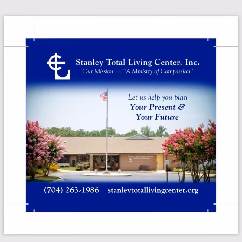 Front exterior view of Stanley Total Living Center building with an American flag on a flagpole in front, surrounded by pink flowering trees and greenery under a clear sky.
