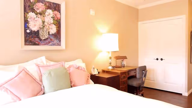 A cozy bedroom with a bed featuring white and pastel pink pillows, a wooden desk with a lamp and chair, beige walls, a white double-door closet, and a floral painting hanging above the bed.