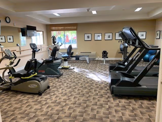 A fitness room with exercise equipment including treadmills, stationary bikes, and a rowing machine. The room has patterned carpet flooring, beige walls with framed pictures, a window with blinds, and a wall-mounted TV.