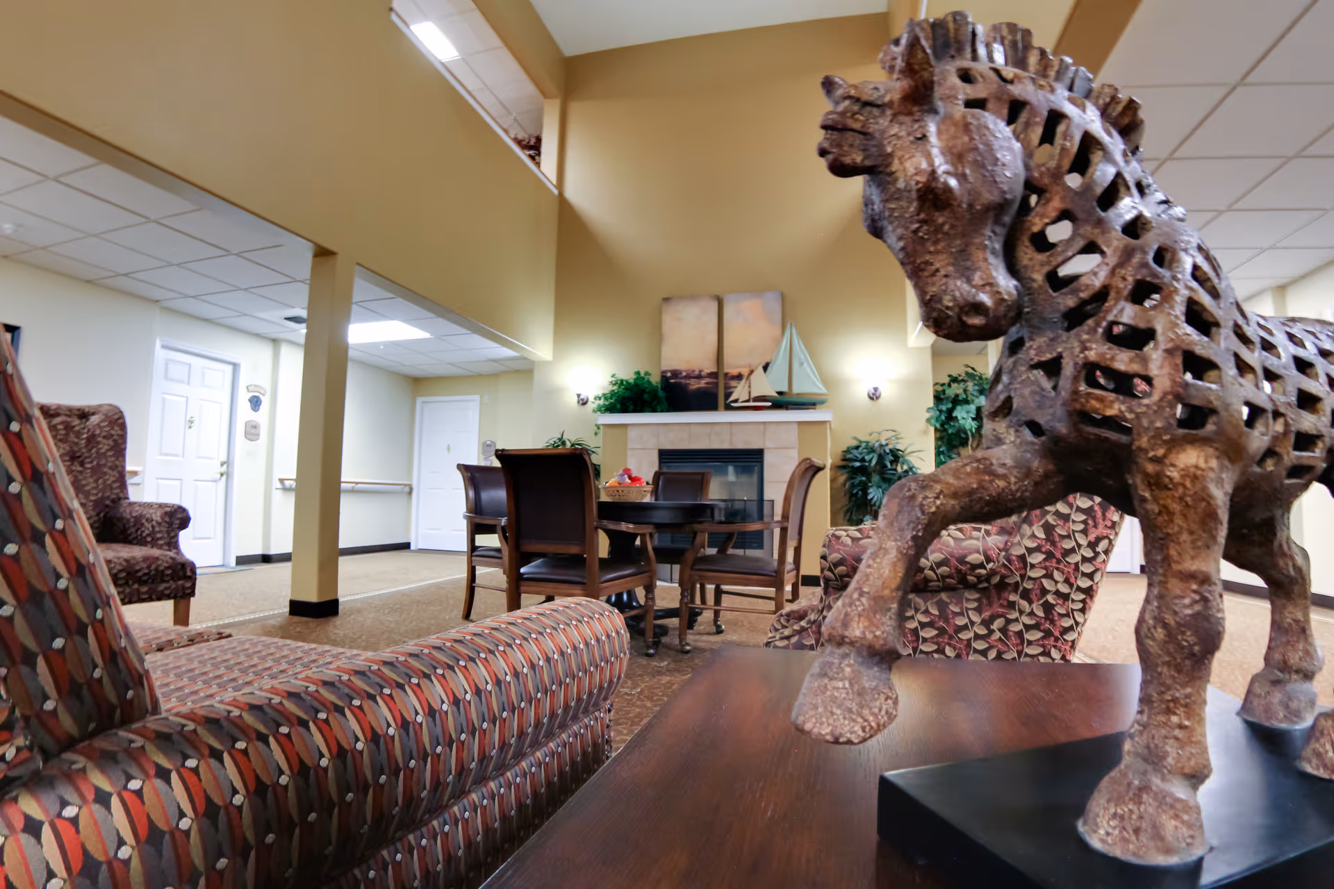 Interior view of a senior living facility lounge area with patterned armchairs, a wooden table with a decorative horse sculpture in the foreground, a round table with chairs near a fireplace, and plants and artwork on the walls.