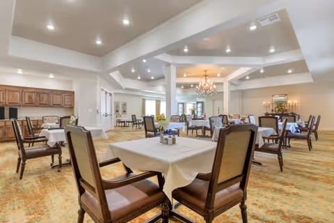 Spacious dining room in a senior living facility with multiple tables covered in white tablecloths and surrounded by wooden chairs with cushioned seats. The room features a beige patterned carpet, recessed lighting in a tray ceiling, and a chandelier hanging in the background. There are decorative elements such as flowers on the tables and framed artwork on the walls.