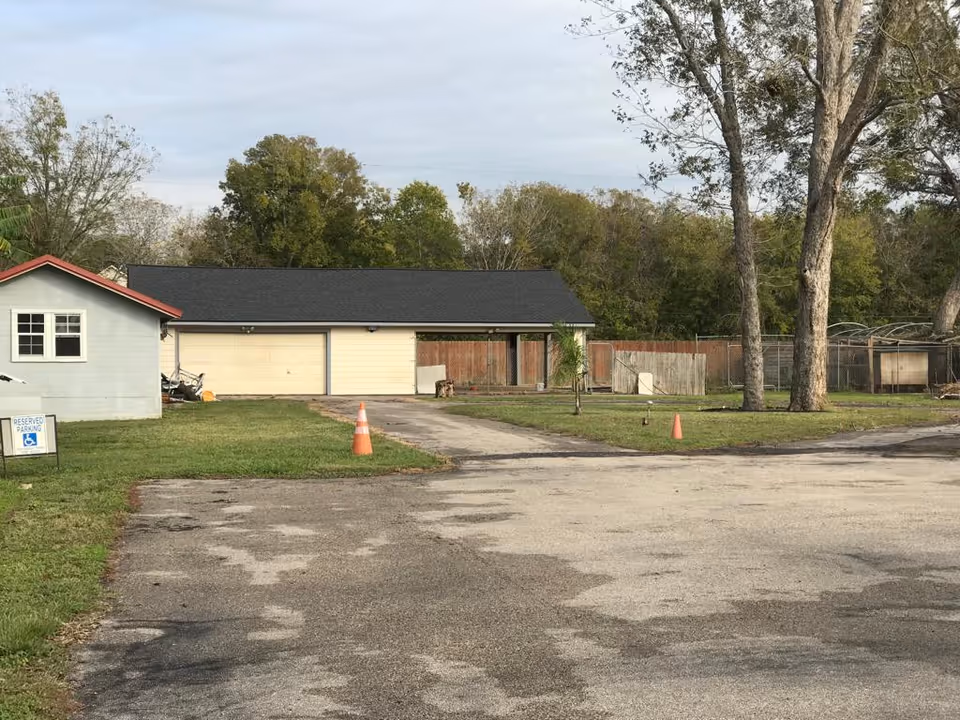 Driveway and parking area leading to a low building with double garage doors, grassy yard, trees, and orange traffic cones.