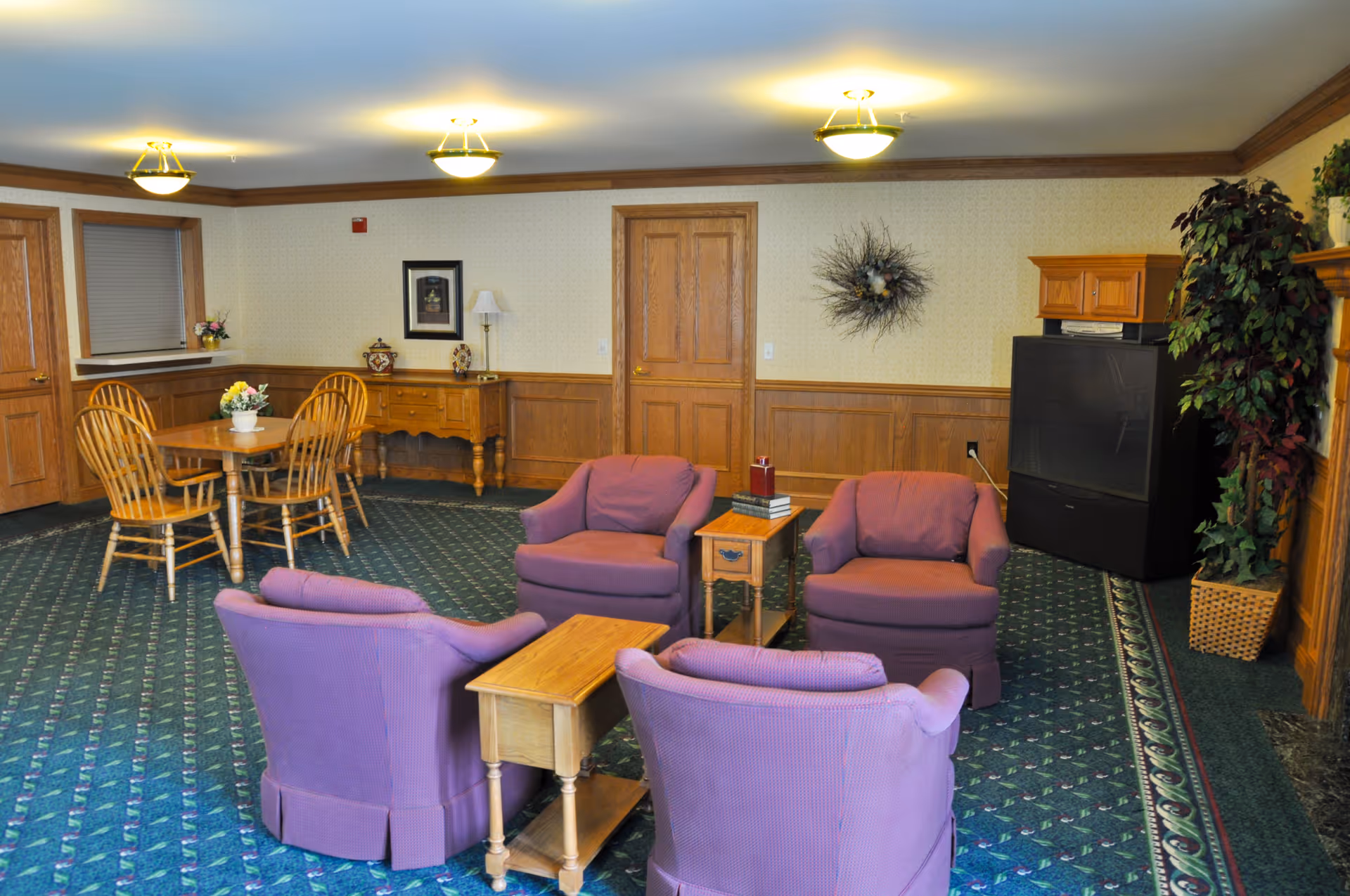 A cozy common area with four purple upholstered armchairs arranged around two small wooden tables. In the background, there is a wooden dining table with four chairs, a sideboard with decorative items, a framed picture, and a wreath on the wall. The room has wood paneling on the lower half of the walls, patterned carpet, and ceiling lights. A large television and a tall potted plant are visible in the corner.