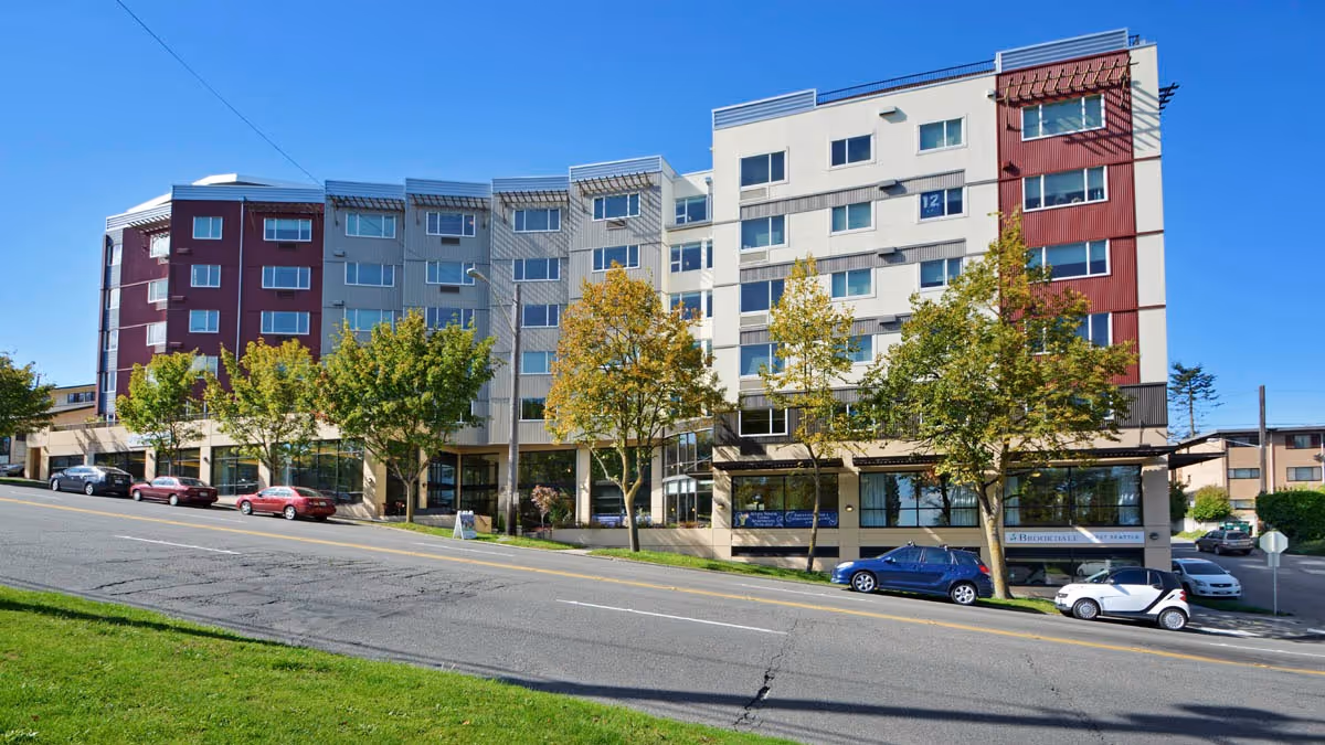 Exterior view of a multi-story senior living facility building with a modern design, featuring large windows and a mix of red and white paneling. Several trees and parked cars line the street in front of the building under a clear blue sky.