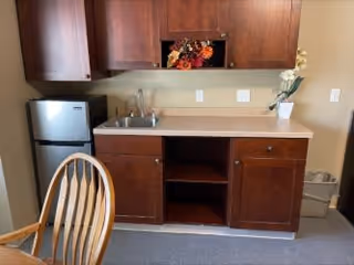A small kitchenette area with dark wooden cabinets, a countertop with a sink, a mini refrigerator, and a wooden chair partially visible in the foreground. There is a decorative floral arrangement in an open cabinet space above the sink and a small vase with white flowers on the countertop. A trash bin is placed on the floor to the right.
