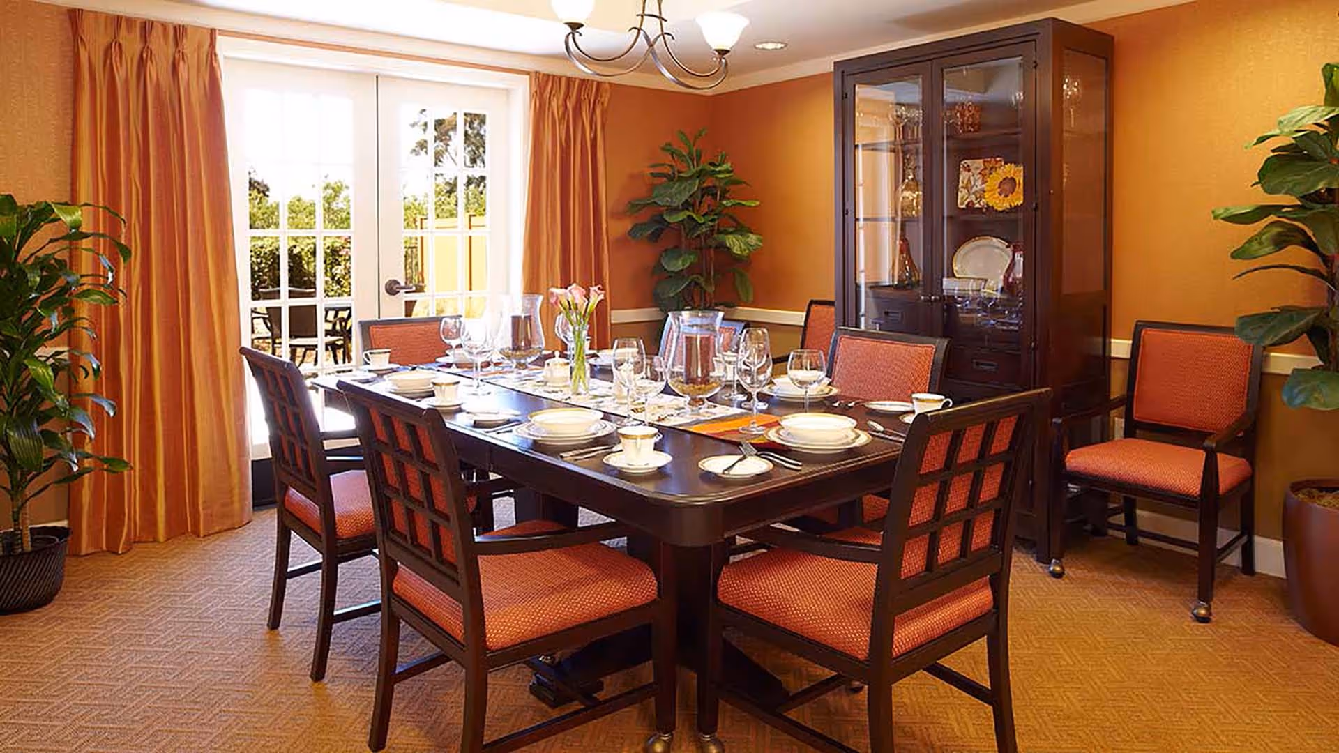 A dining room with a large rectangular wooden table set for a meal with plates, bowls, cups, and glassware. The room features orange walls, matching orange curtains, and six wooden chairs with orange cushions around the table. There are two large potted plants and a dark wooden china cabinet displaying decorative plates and glassware. French doors with glass panes lead outside to a patio area.