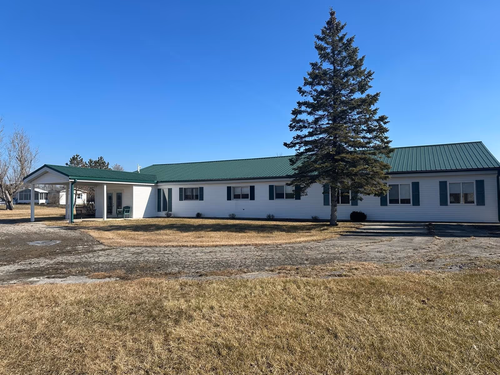 Single-story white building with a green metal roof and green shutters, featuring a covered entrance with two chairs outside. A large evergreen tree stands in front of the building on a dry grassy area under a clear blue sky.