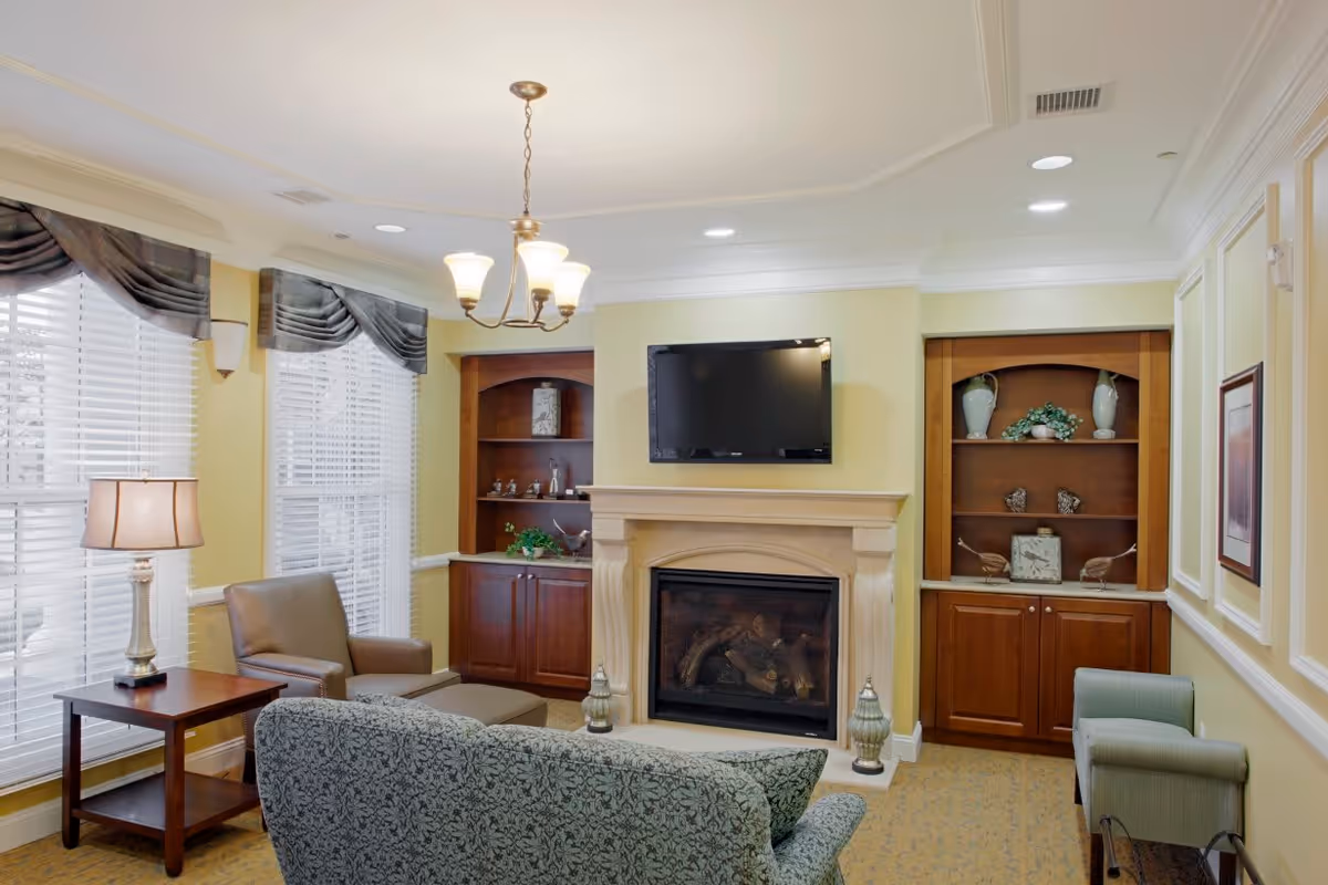A cozy living room with a patterned sofa and armchair facing a fireplace with a mounted flat-screen TV above it. The room has built-in wooden shelves on either side of the fireplace, decorated with vases and small ornaments. Large windows with blinds and valances let in natural light, and a table lamp sits on a side table next to the armchair. The walls are painted light yellow with white trim, and a chandelier hangs from the ceiling.