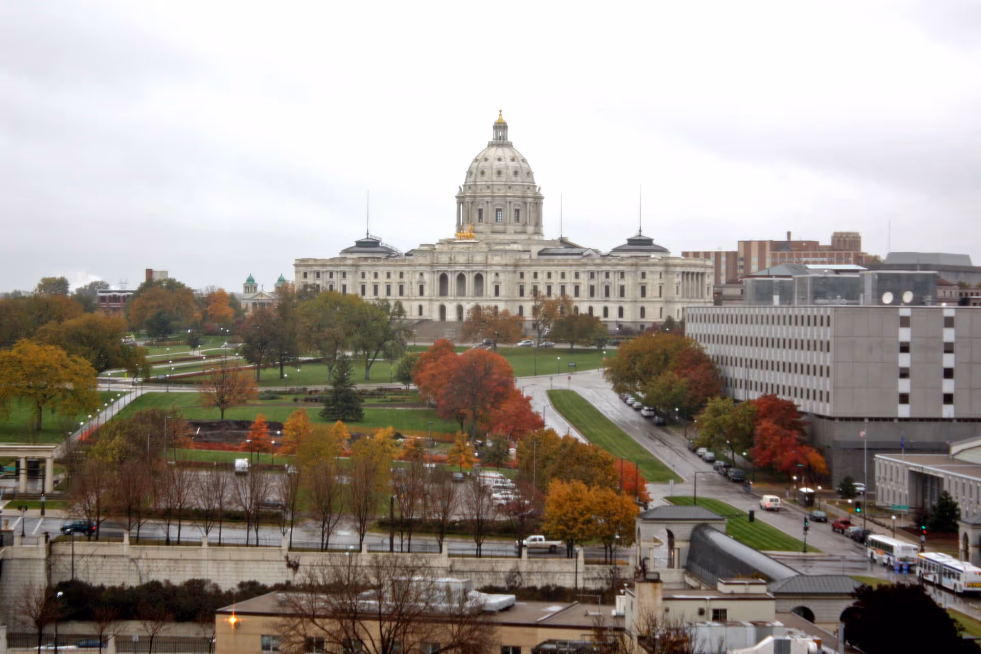 View of a large classical government building with a dome, surrounded by trees with autumn foliage and adjacent city streets on a cloudy day.