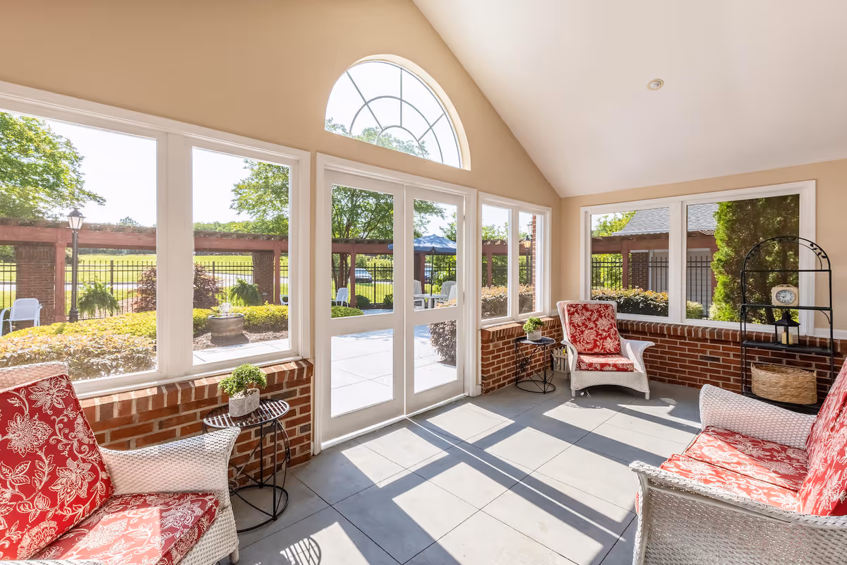 A bright sunroom with large windows and a glass door leading outside. The room features white wicker furniture with red floral cushions, small round side tables with potted plants, and a black metal shelving unit with decorative items. Outside, there is a garden area with greenery, a brick pergola, and outdoor seating.