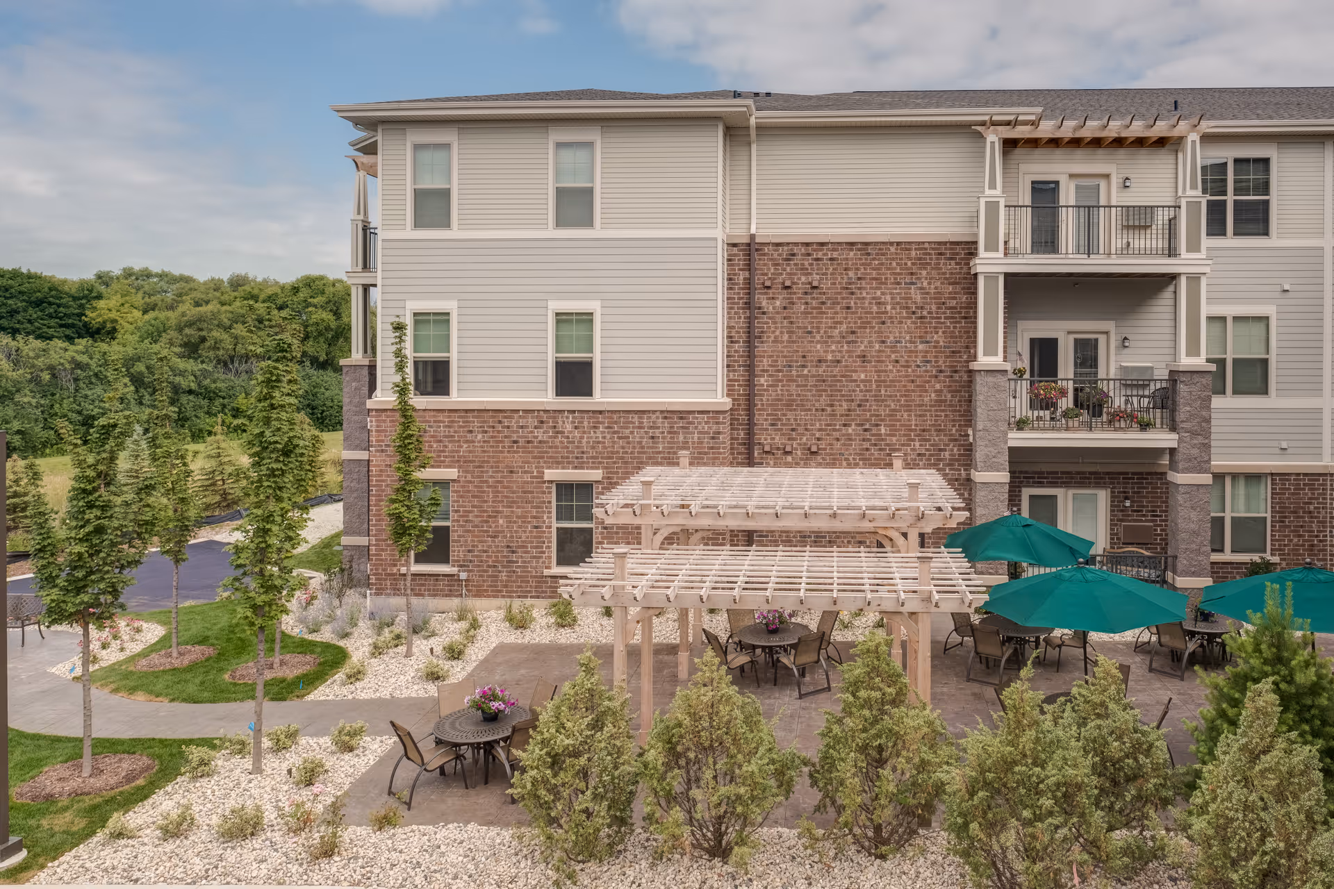 Outdoor patio area at Heritage Lake Country Senior Living featuring multiple tables with chairs, some under green umbrellas and others under wooden pergolas. The patio is surrounded by small trees, shrubs, and landscaped rock beds. The background shows a multi-story building with brick and siding exterior and balconies with potted plants.