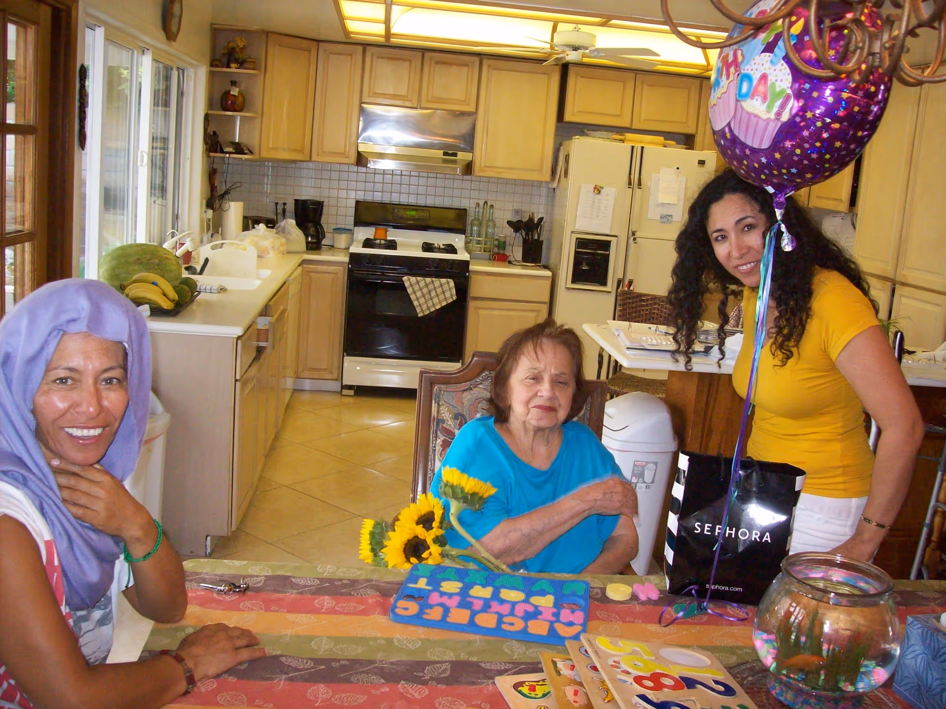 Three women in a kitchen setting. One woman on the left is smiling and wearing a purple headscarf. An elderly woman in the center is seated at a table with a colorful alphabet puzzle and sunflowers in front of her. Another woman on the right, wearing a yellow shirt, is standing and holding a purple birthday balloon. The kitchen has light wood cabinets, a white refrigerator, and a stove in the background.