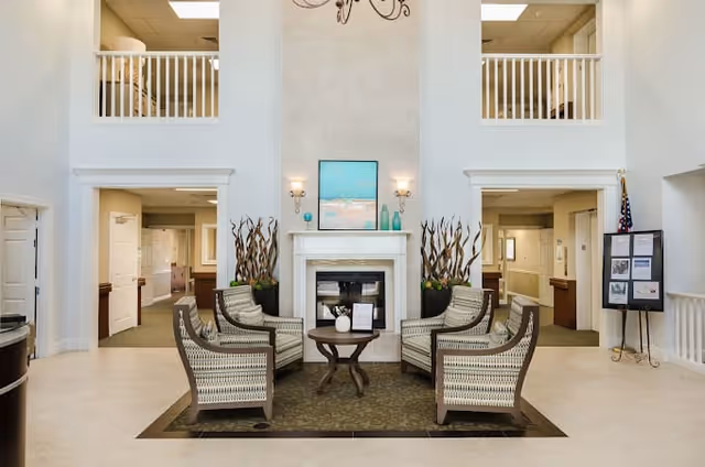 Two-story lobby seating area with four chairs arranged around a small table in front of a fireplace beneath a mezzanine with railings.
