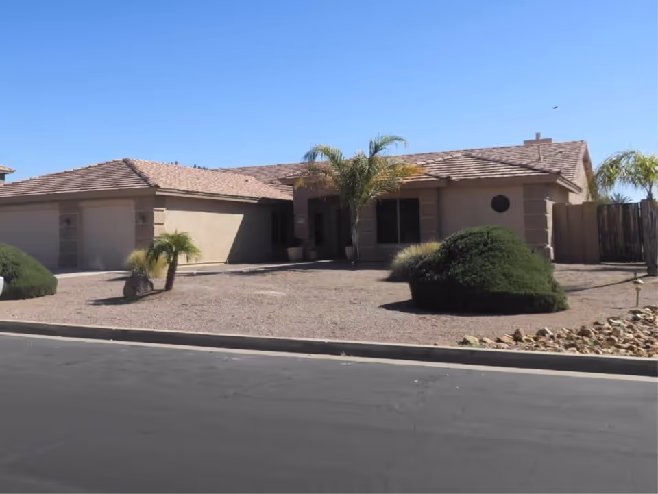 Single-story residential building with a tiled roof, beige exterior walls, and a two-car garage. The front yard features desert landscaping with gravel, small palm trees, and trimmed bushes under a clear blue sky.