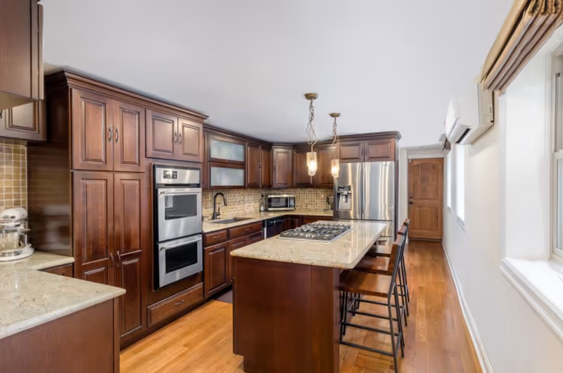 A modern kitchen with dark wooden cabinets, granite countertops, a central island with a built-in gas stove, two hanging pendant lights, stainless steel double oven, refrigerator, and wooden flooring. There are two high chairs at the island and a wooden door at the far end of the room.