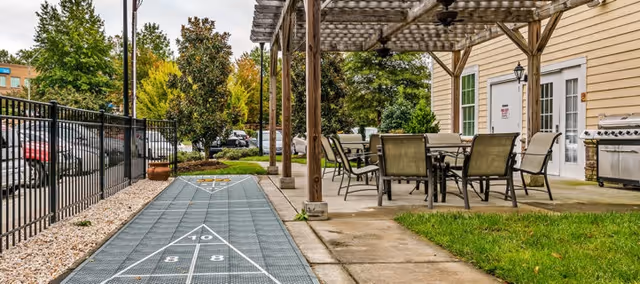 Outdoor patio area at Cadence at Wake Forest featuring a shuffleboard court on the left, a covered seating area with tables and chairs on the right, a barbecue grill near the building, and trees and parked cars in the background.