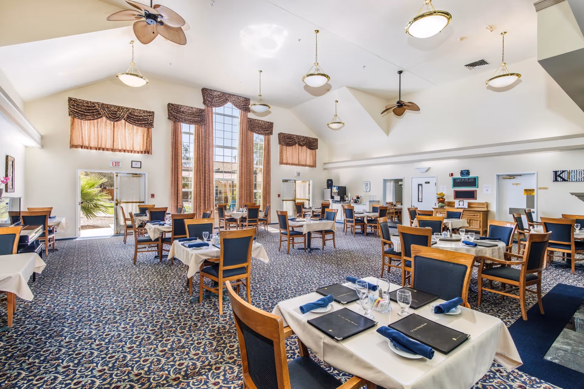 A spacious dining room with multiple tables covered in white tablecloths, each set with menus, glasses, plates, and blue napkins. The room features large windows with brown curtains, ceiling fans, and hanging light fixtures. The carpet has a blue and beige pattern, and there are exit doors visible in the background.