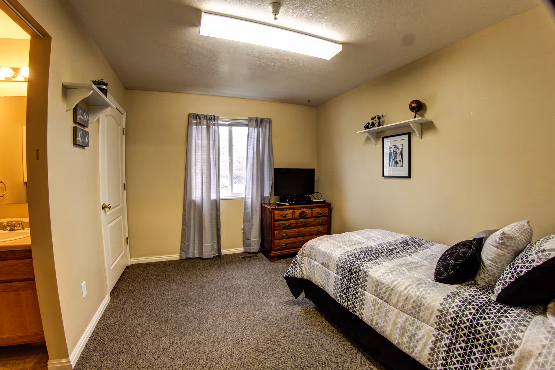 A small furnished bedroom with a bed, dresser and TV, a window with curtains, and a doorway to a bathroom.
