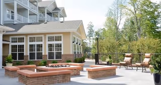 Outdoor patio area at a senior living facility with brick fire pit surrounded by built-in brick seating, two cushioned chairs, potted plants, and a multi-story building with large windows in the background.