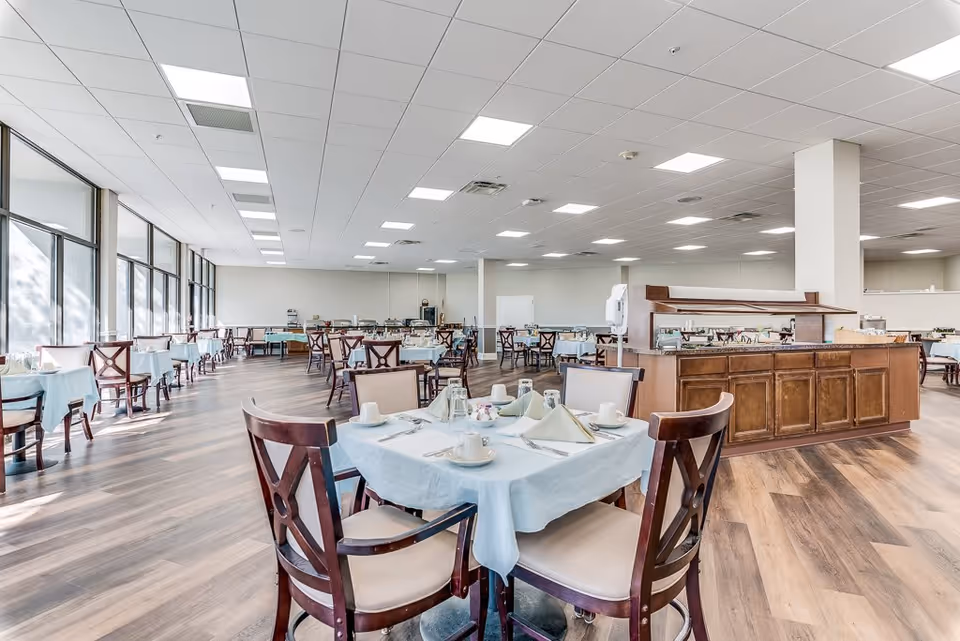 Spacious dining room with multiple tables set with tablecloths and place settings, large windows along one side and a central buffet station.