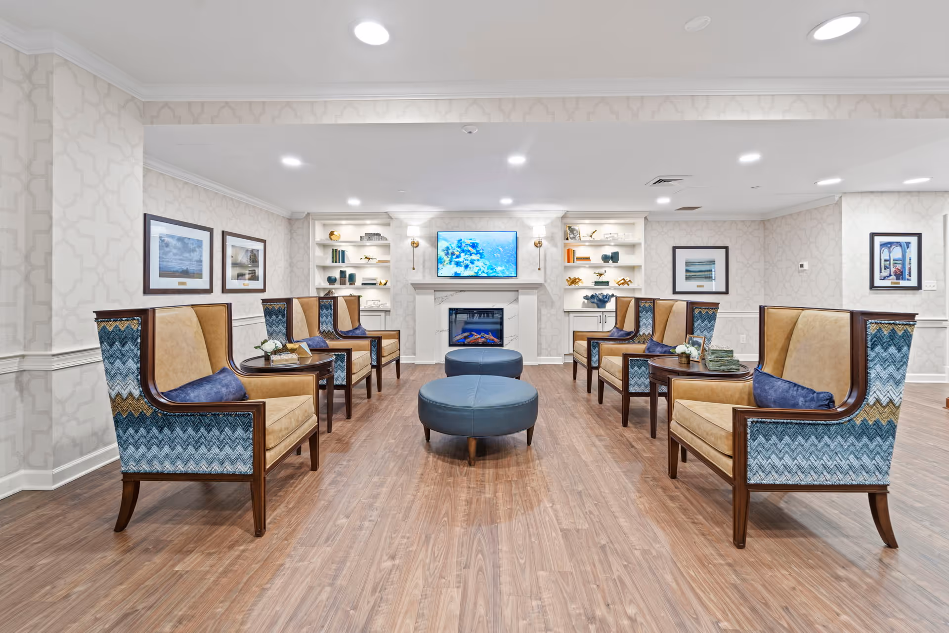 A bright and spacious senior living facility common area with wooden flooring, patterned wallpaper, and recessed ceiling lights. The room features two rows of high-backed armchairs with blue and tan upholstery facing each other, separated by round wooden side tables with decorative items. At the far end, there is a white fireplace with a flat-screen TV mounted above it, flanked by built-in shelves displaying books and decorative objects. Framed artwork is hung on the walls.