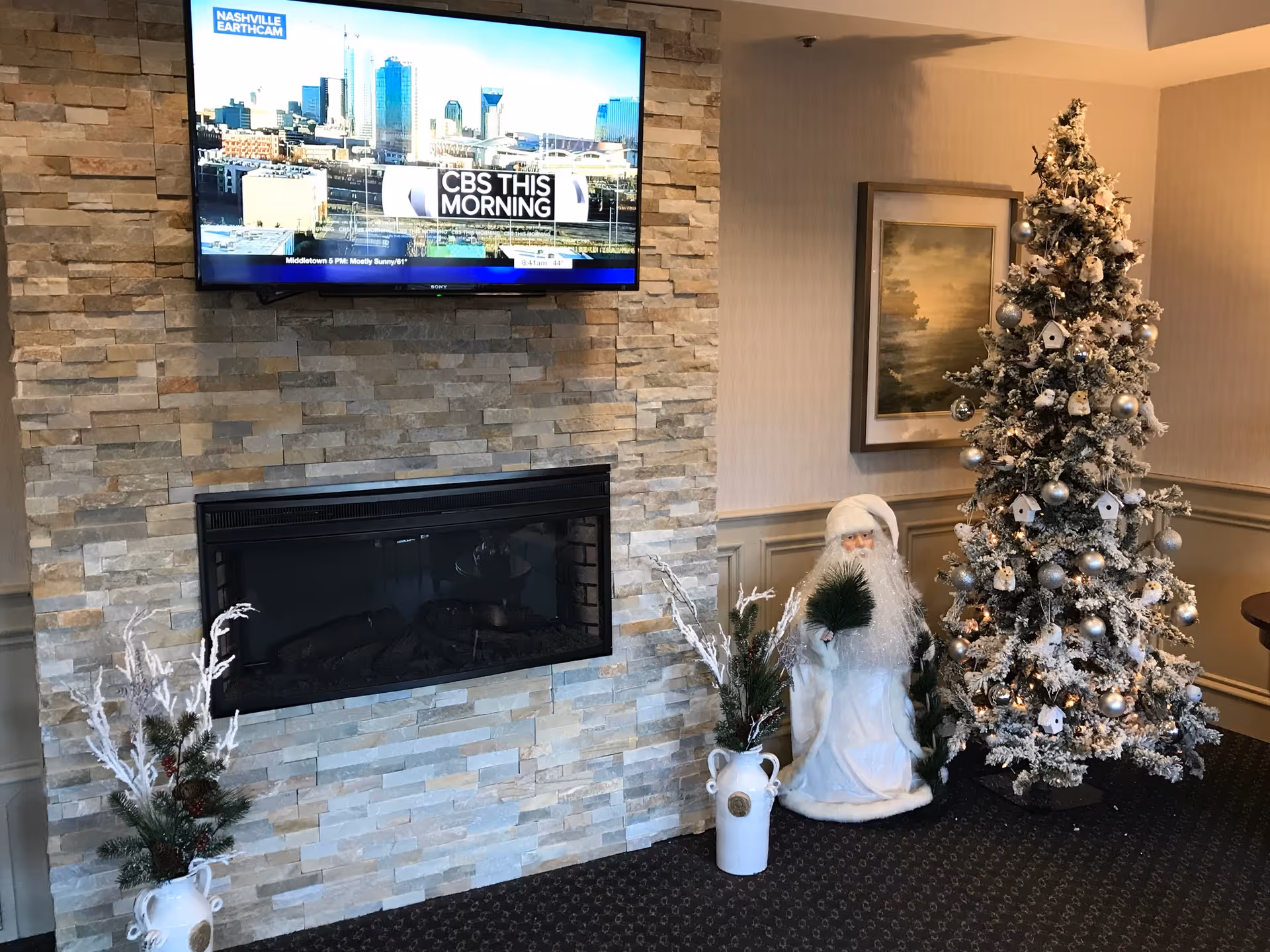 A cozy interior corner featuring a stone fireplace with a mounted flat-screen TV displaying CBS This Morning. To the right of the fireplace is a decorated Christmas tree with silver ornaments and small white birdhouses, a white Santa Claus figure holding greenery, and two white vases with festive greenery and white branches. A framed landscape painting hangs on the wall behind the decorations.