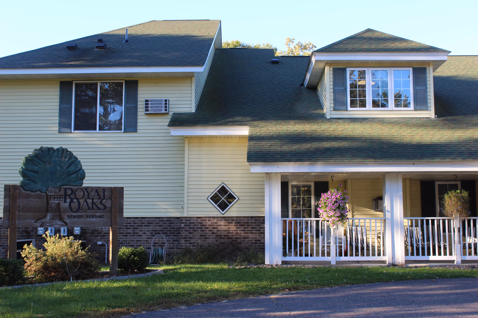 Exterior view of a senior housing building with yellow siding, green roof shingles, and white trim. There is a porch with white railings and hanging flower baskets. A wooden sign in front reads 'The Royal Oaks Senior Housing'.