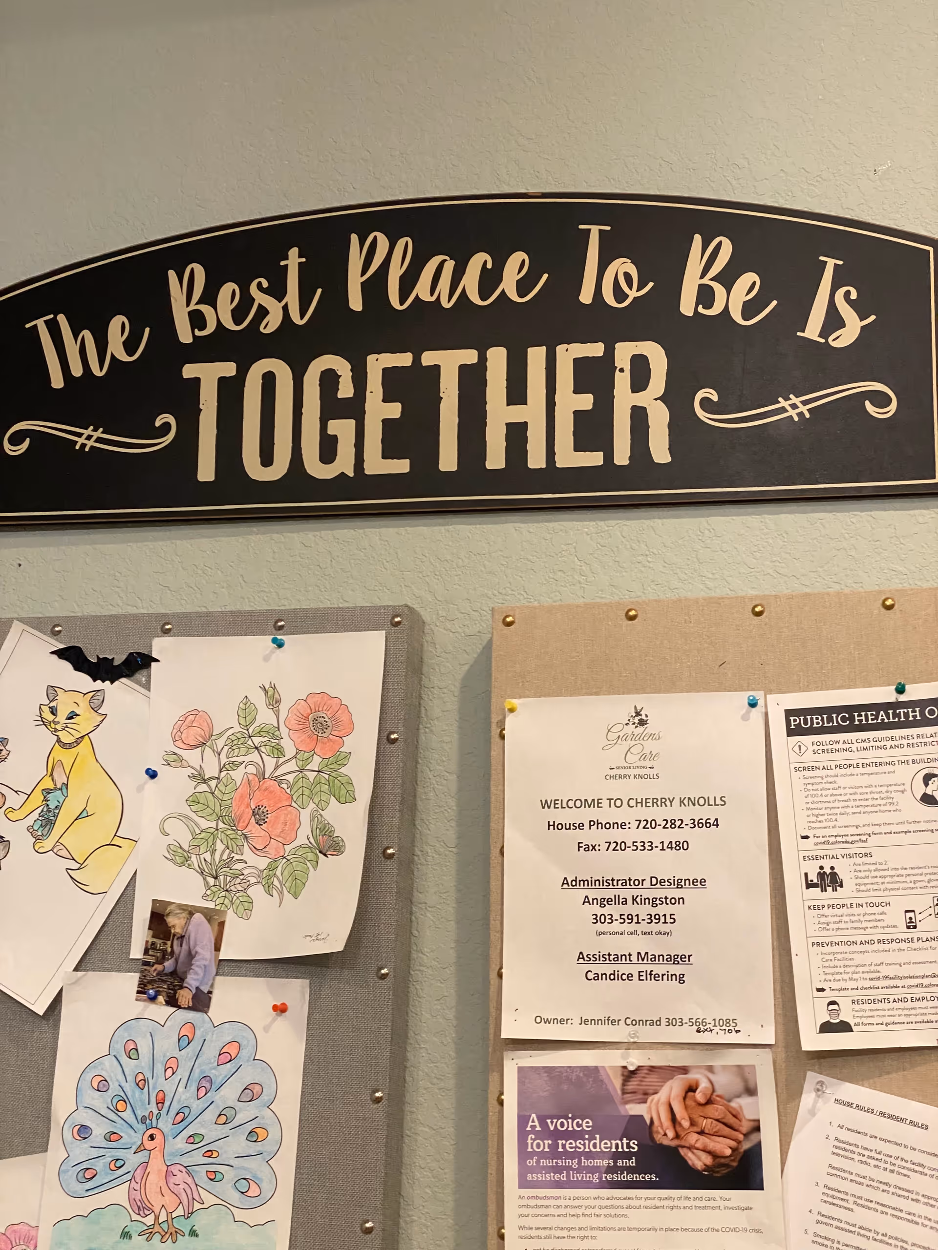 A bulletin board with various pinned papers and drawings, including a colored drawing of a cat, flowers, and a peacock. Above the board is a black sign with white text that reads 'The Best Place To Be Is TOGETHER.'