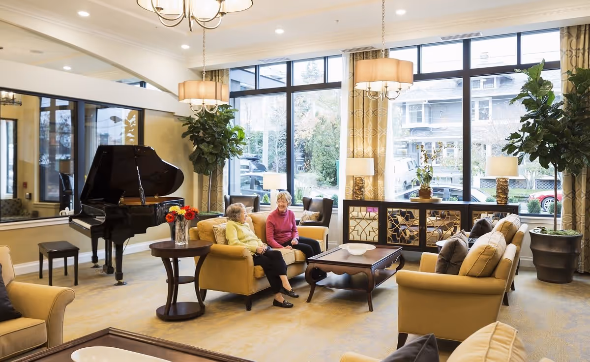 A bright and spacious living room area with large windows letting in natural light. Two elderly women are sitting and chatting on a beige sofa. The room features comfortable beige armchairs, a grand piano, side tables with lamps, a decorative cabinet, and large potted plants. The decor includes patterned curtains and a chandelier overhead.