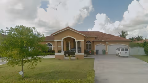 Single-story tan retirement home with a front entrance, driveway, and parked vehicles under a cloudy sky.