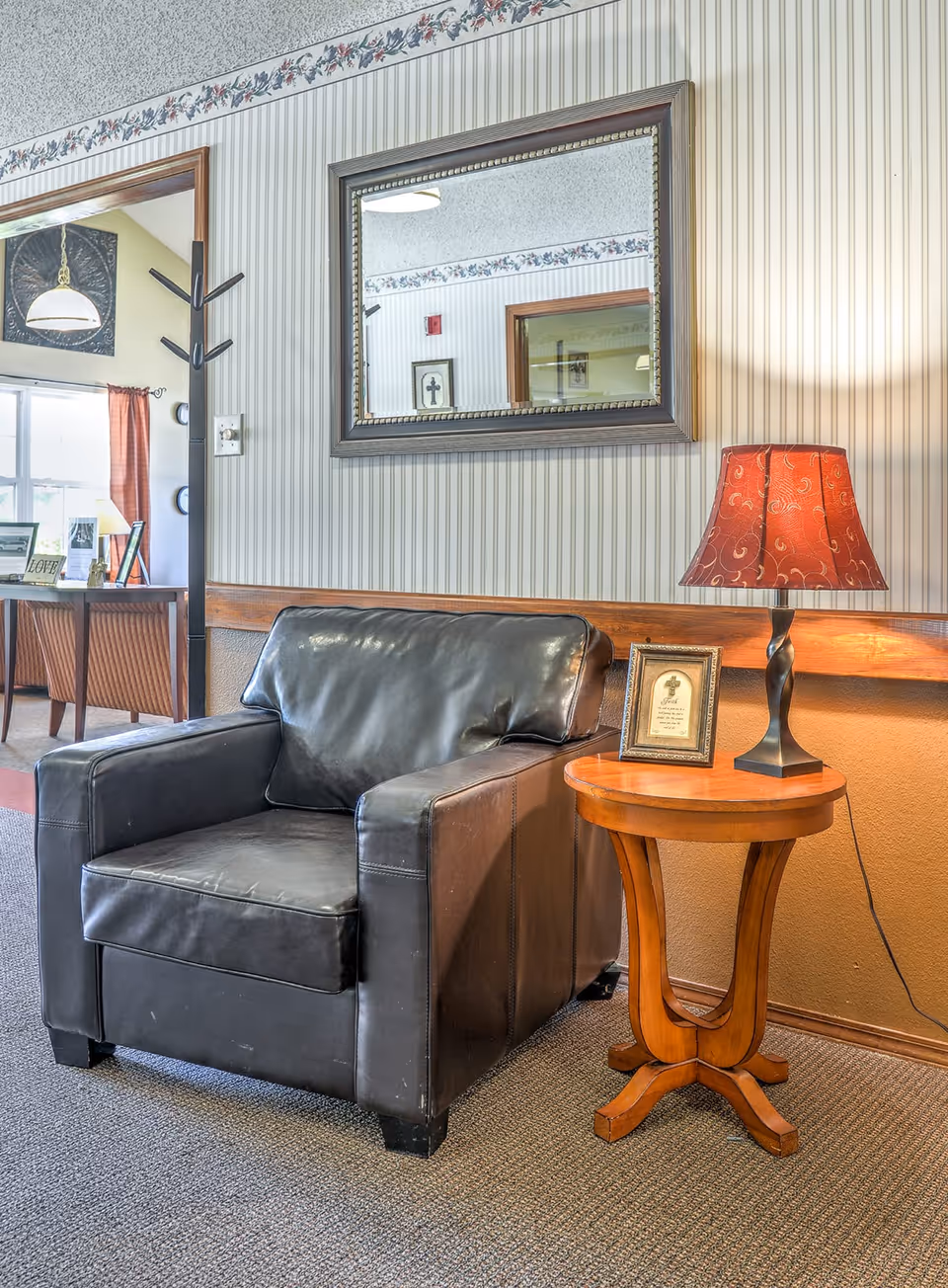 A cozy seating area in a senior living facility featuring a black leather armchair next to a wooden side table with a red patterned lampshade and a framed picture. A large mirror hangs on the striped wallpapered wall above the chair, reflecting part of the room including a doorway and framed artwork.