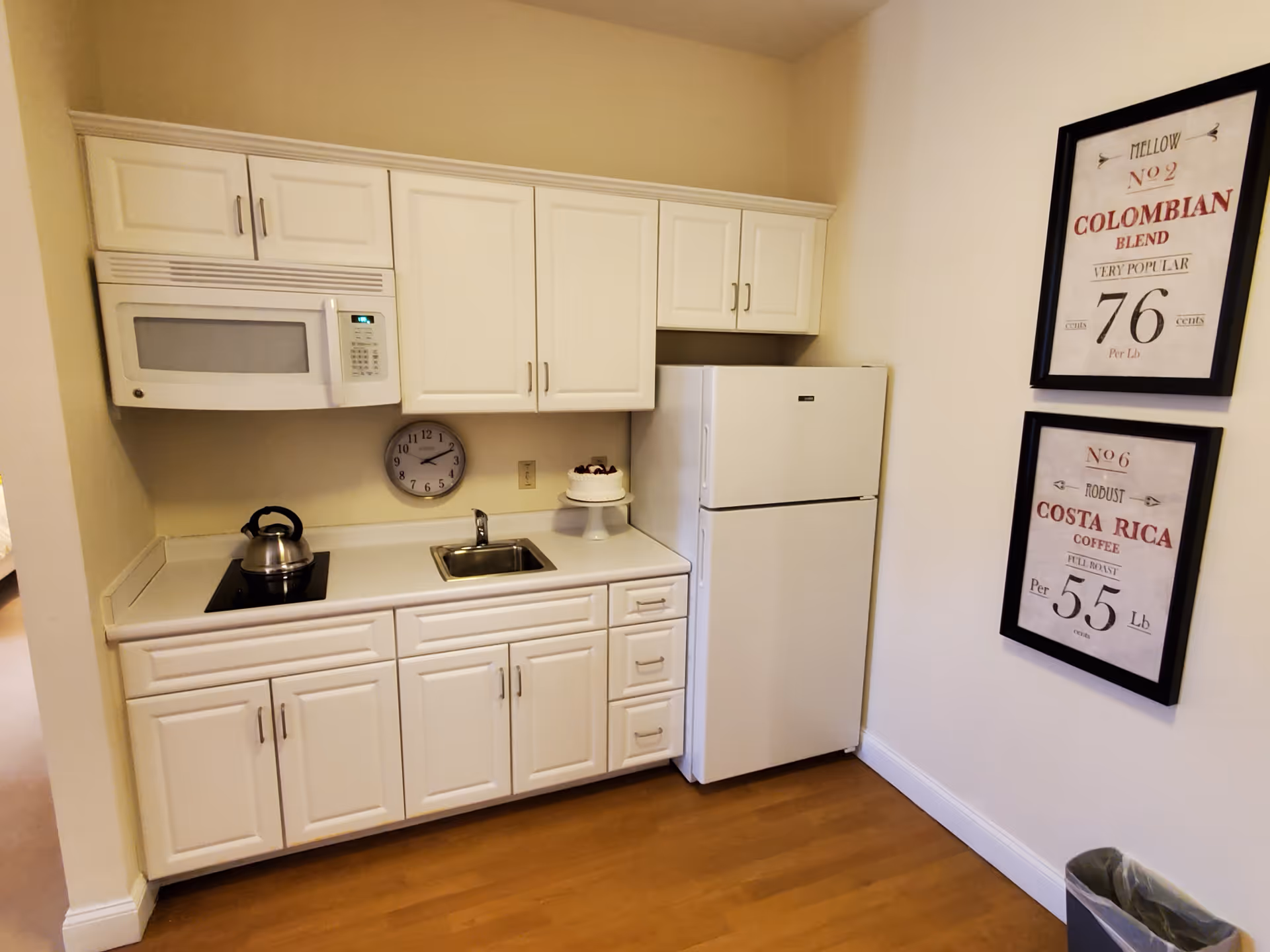 A small kitchen area with white cabinets, a microwave, a stovetop with a kettle, a sink, and a refrigerator. There are two framed coffee-themed prints on the adjacent wall and a small cake on a stand on the counter. The floor is wooden.