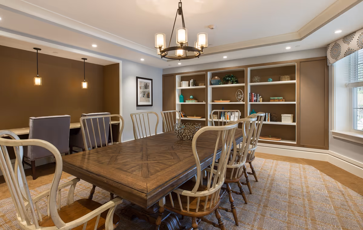 A bright dining room with a large wooden table surrounded by chairs, built-in shelving, pendant lights and a window.