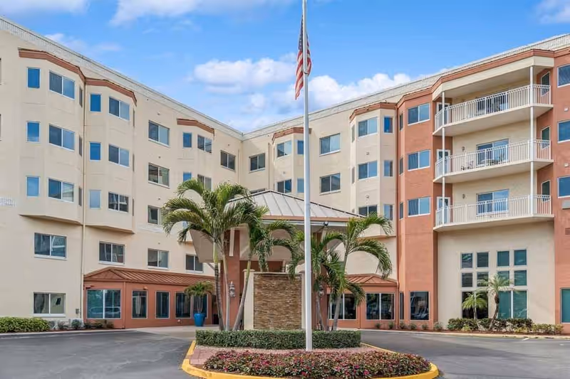 Exterior view of a multi-story senior living facility building with beige and terracotta colored walls, multiple windows, balconies, palm trees, and an American flag on a flagpole in front of the entrance.