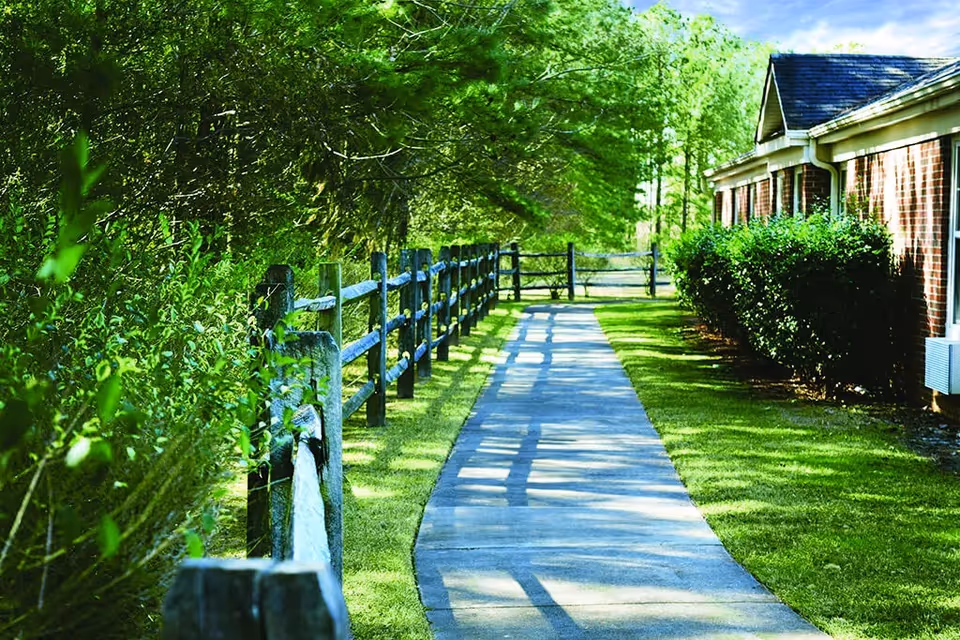 A paved walkway runs between a wooden split-rail fence and a brick building lined with shrubs beneath leafy trees.