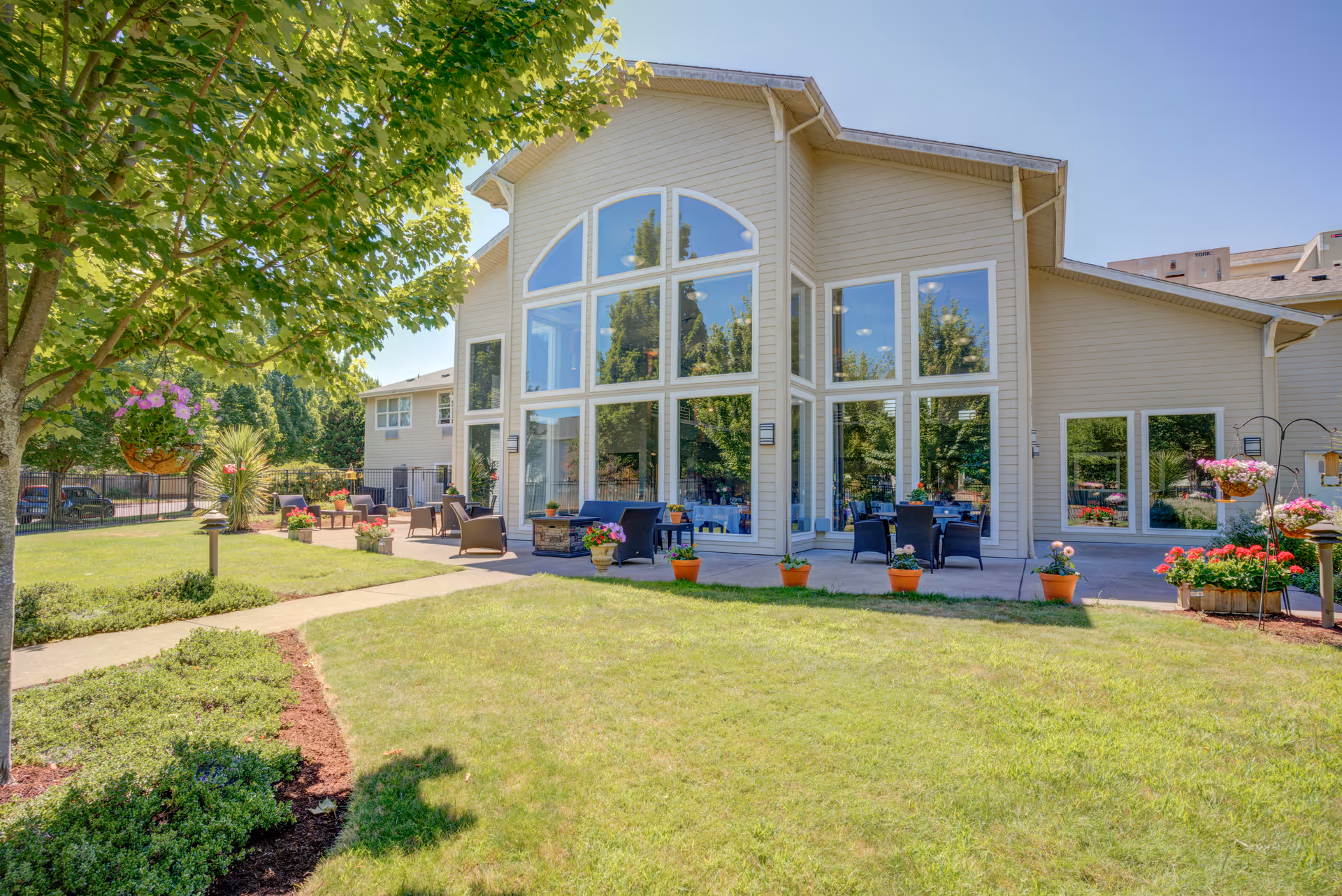 Exterior front of a senior living facility with large arched windows, patio seating, potted flowers, and a grassy courtyard.