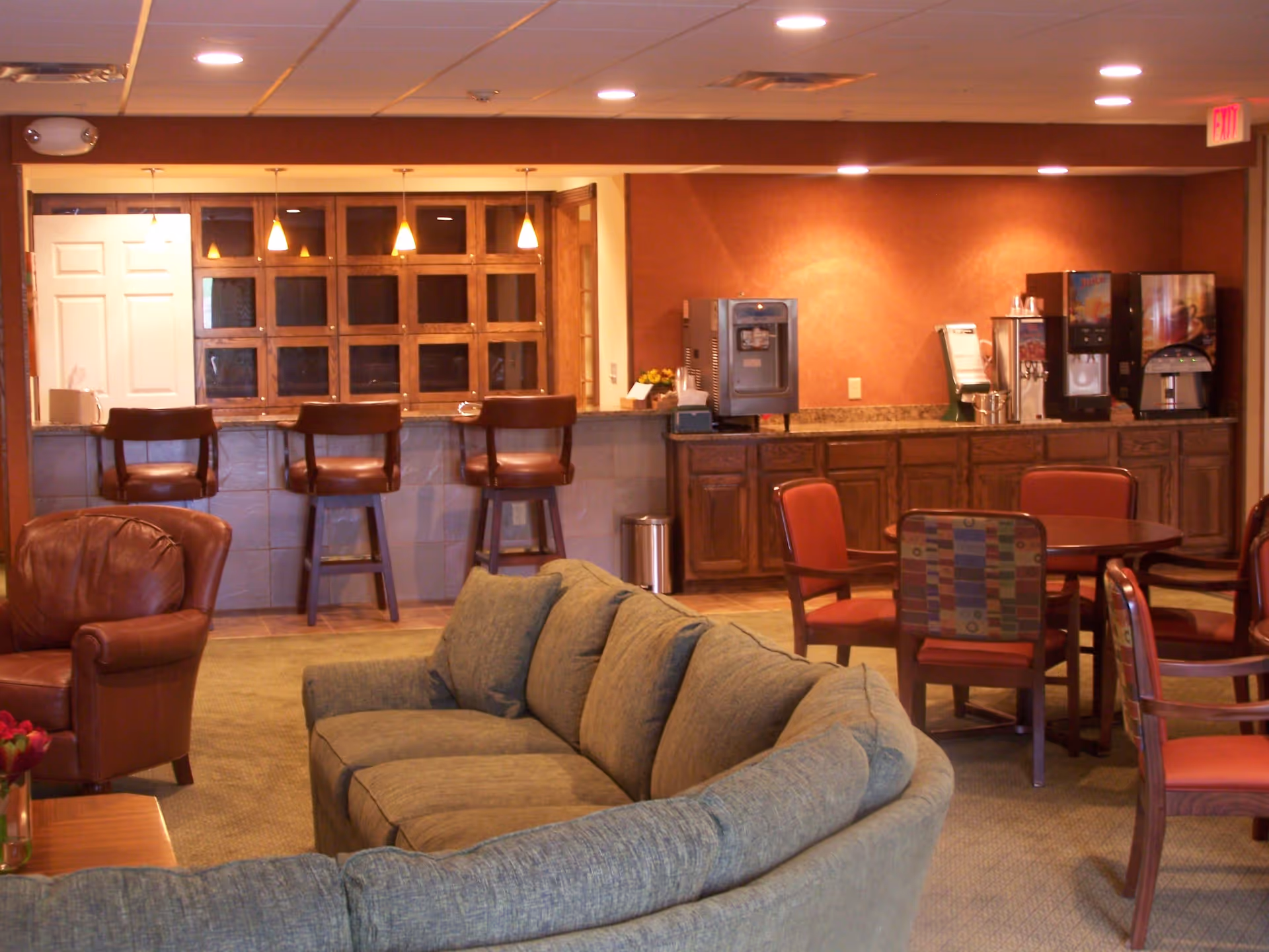 A cozy common area in a retirement community featuring a curved gray sofa, a brown leather armchair, a round table with red cushioned chairs, a counter with three bar stools, and a beverage station with coffee and drink machines against a warm-toned wall.