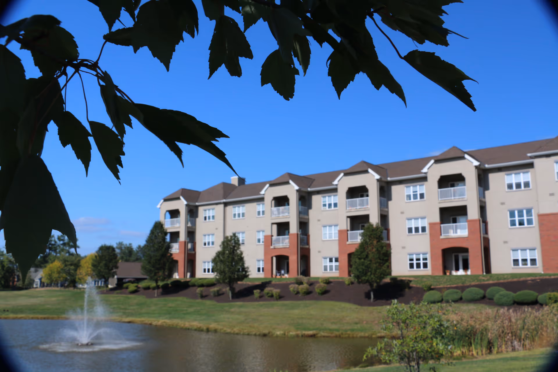 A multi-story residential building with balconies overlooking a pond with a water fountain. The building is surrounded by well-maintained landscaping including trees, bushes, and grass. The sky is clear and blue, and tree leaves are visible in the foreground at the top of the image.