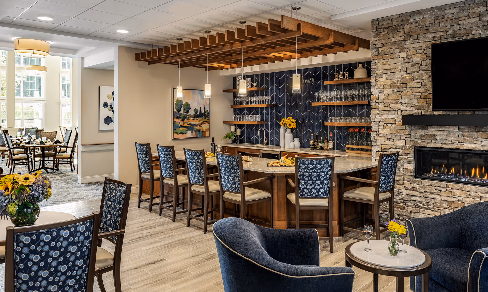 A cozy and elegant common area in a retirement community featuring a bar with high chairs, a stone fireplace with a mounted TV above it, and comfortable seating with small tables decorated with flowers. The background shows a dining area with tables and chairs near large windows letting in natural light.