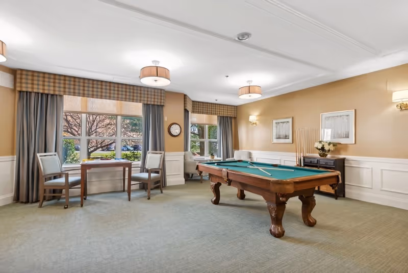 A well-lit recreational room with a pool table in the center, two wooden chairs and a small table near large windows with curtains, a clock on the wall, and a sideboard with flowers and framed pictures on the wall.