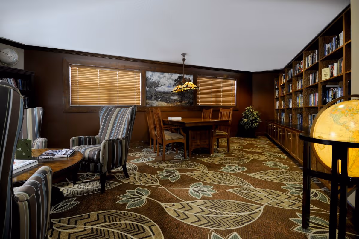 A cozy interior room with a patterned carpet featuring large floral designs. The room has two windows with wooden blinds, a wooden table with six chairs, and a hanging light fixture above the table. There are two striped armchairs around a small round wooden table with books on it. A large bookshelf filled with books lines one wall, and a globe on a stand is visible in the foreground. A potted plant is placed in the corner near the bookshelf.
