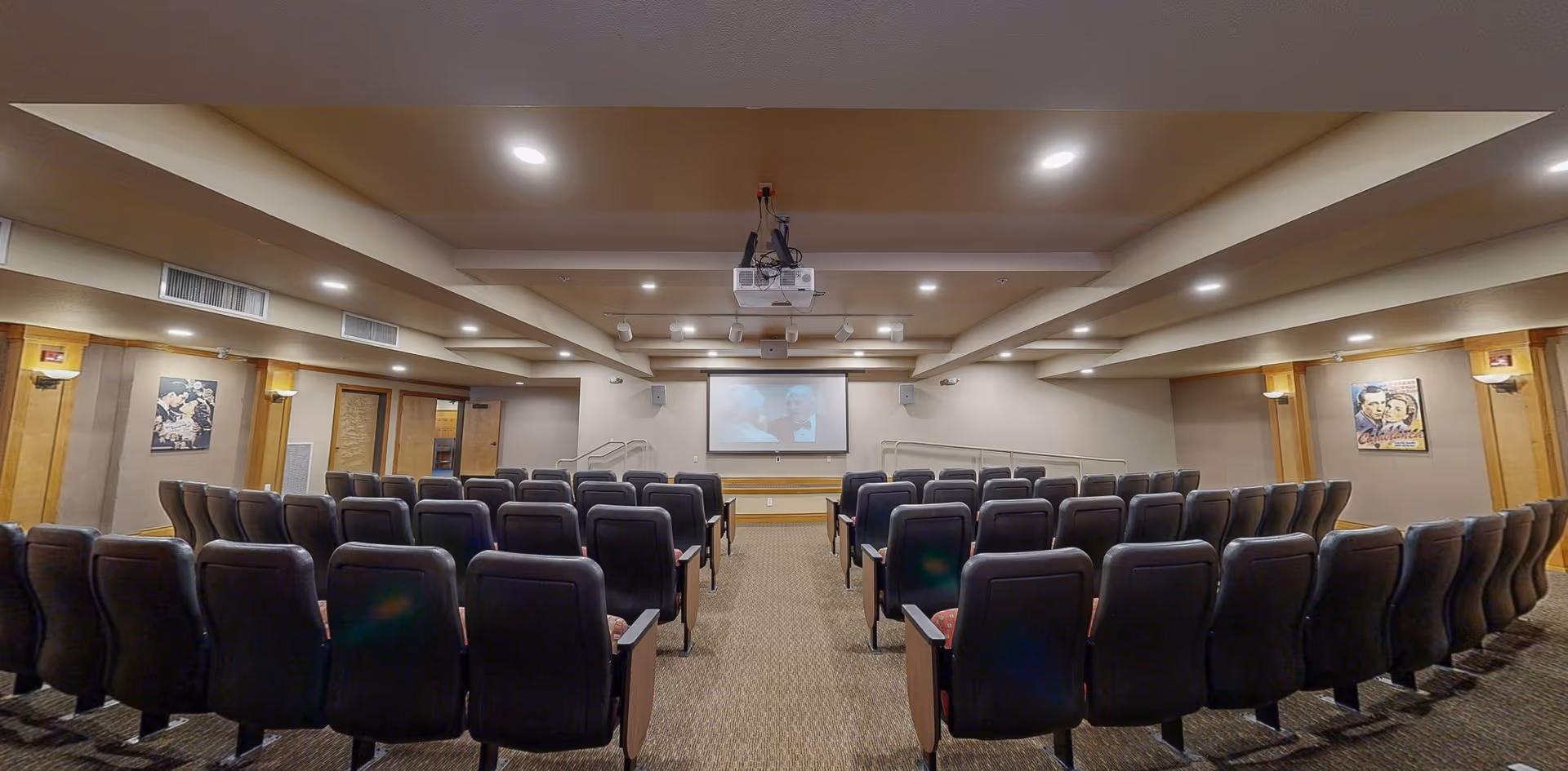 Small theater-style screening room with rows of upholstered seats facing a projection screen and a ceiling-mounted projector.