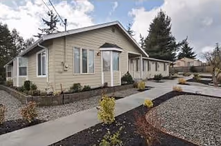 Single-story beige residential building with a paved walkway and landscaped gravel beds under a partly cloudy sky.