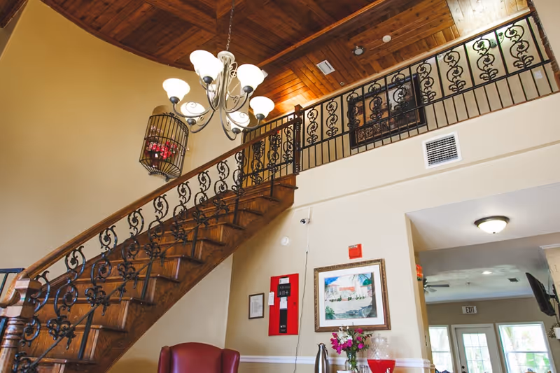 Interior view of a senior living facility showing a wooden staircase with ornate black metal railings leading to an upper floor. A chandelier with multiple lights hangs from a wooden ceiling. Below the staircase is a red armchair, a framed painting on the wall, a red fire alarm panel, and a table with flowers and a beverage dispenser. The area is well-lit with natural light coming through windows and doors in the background.
