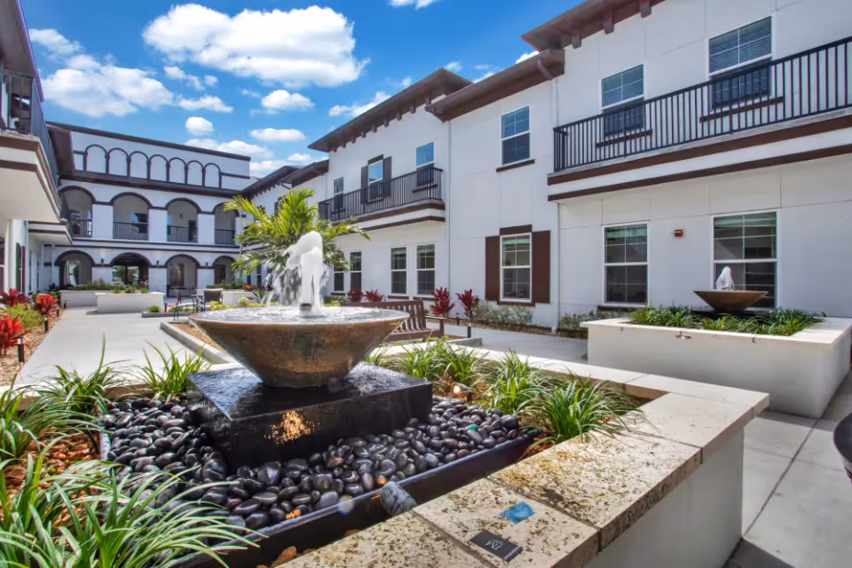 Outdoor courtyard area of The Blake at St. Johns facility featuring a central water fountain surrounded by black stones and greenery, with benches and landscaped plants along the walkway. The building has white walls with brown trim and balconies, under a blue sky with scattered clouds.