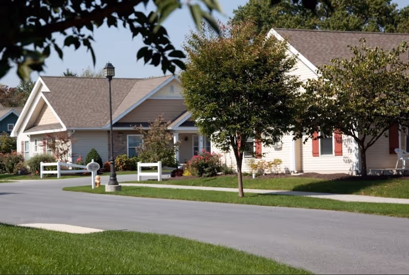 A quiet residential street in a senior living community with single-story houses, trees, and well-maintained lawns under a clear sky.