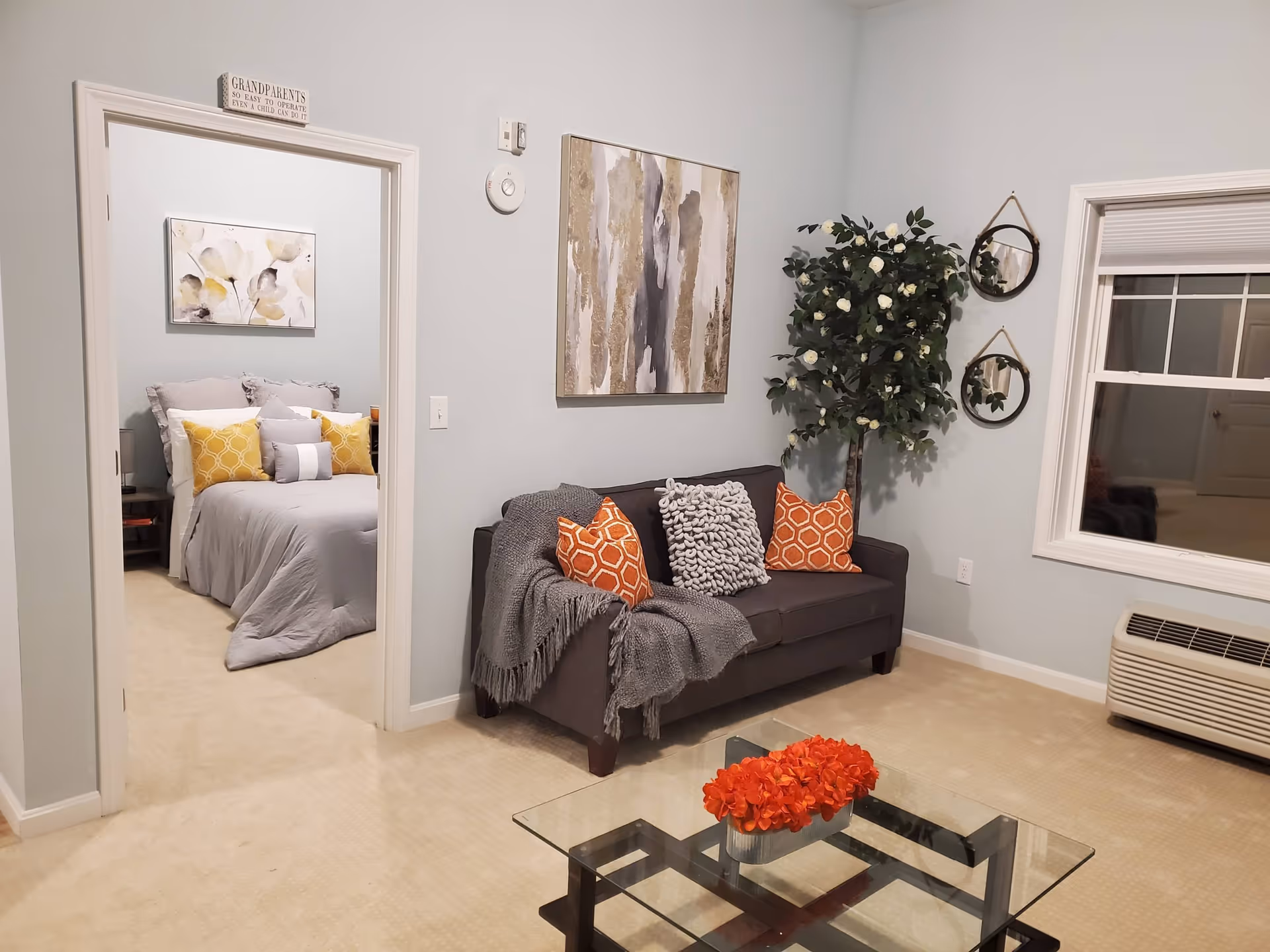 A cozy living room with a dark gray sofa adorned with orange and gray pillows and a gray throw blanket. A glass coffee table with a red floral centerpiece is in front of the sofa. To the right, there is a window with a white frame and an air conditioning unit below it. A tall artificial plant with white flowers stands in the corner next to two round hanging mirrors. Through an open doorway, a bedroom is visible with a neatly made bed featuring gray bedding and yellow accent pillows, and a floral painting above the bed. A sign above the doorway reads 'GRANDPARENTS SO EASY TO OPERATE EVEN A CHILD CAN DO IT.' The walls are painted light gray and the floor is carpeted in beige.