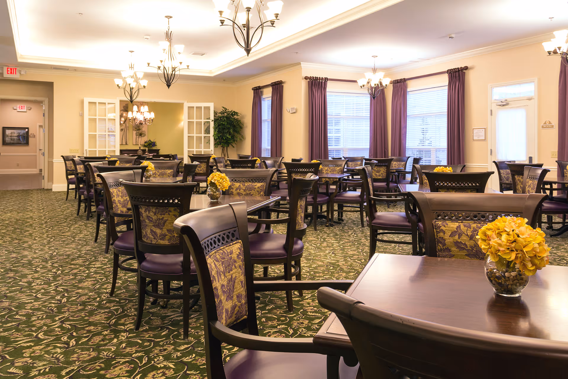 A spacious dining room with multiple dark wood tables and chairs featuring purple and floral upholstery. Each table has a small vase with yellow flowers. The room has large windows with purple curtains, green patterned carpet, and several chandeliers hanging from the ceiling providing warm lighting.